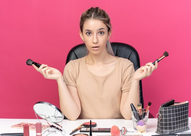 Hermosa joven sorprendida se sienta a la mesa con herramientas de maquillaje sosteniendo pincel en polvo extendiendo las manos aisladas sobre fondo rosa