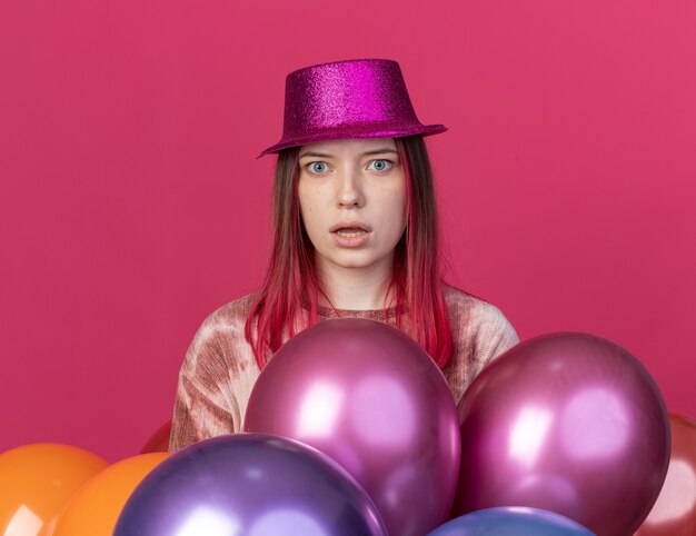 Hermosa joven sorprendida con sombrero de fiesta de pie detrás de globos aislados en la pared rosa
