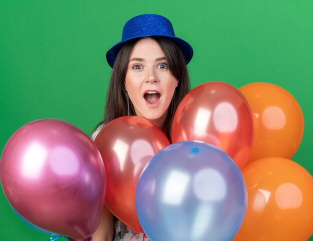 Hermosa joven sorprendida con sombrero de fiesta de pie detrás de globos aislados en la pared verde