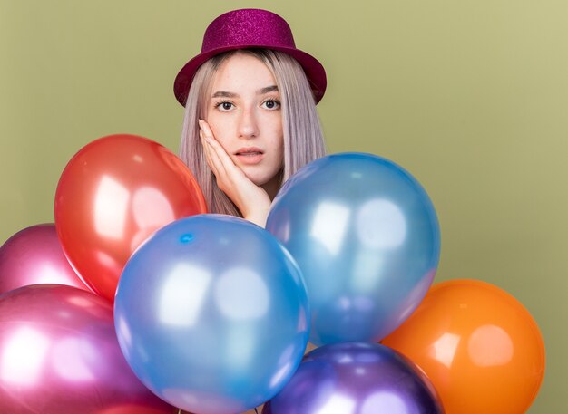 Hermosa joven sorprendida con sombrero de fiesta de pie detrás de globos poniendo la mano en la mejilla