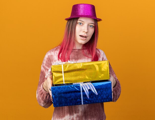 Hermosa joven sorprendida con sombrero de fiesta sosteniendo cajas de regalo aisladas en la pared naranja