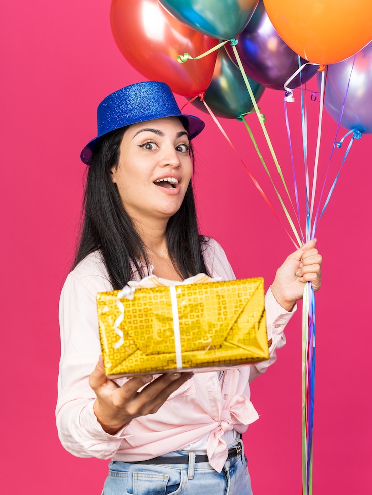 Foto gratuito hermosa joven sorprendida con sombrero de fiesta sosteniendo globos con caja de regalo aislada en la pared rosa