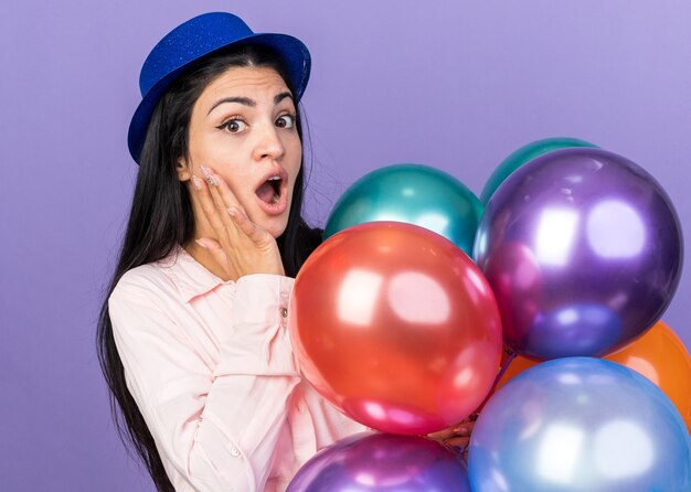 Hermosa joven sorprendida con sombrero de fiesta sosteniendo globos poniendo la mano en la mejilla aislada en la pared azul