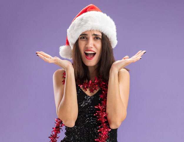 Hermosa joven sorprendida con sombrero de navidad con guirnalda en el cuello extendiendo las manos aisladas sobre fondo púrpura