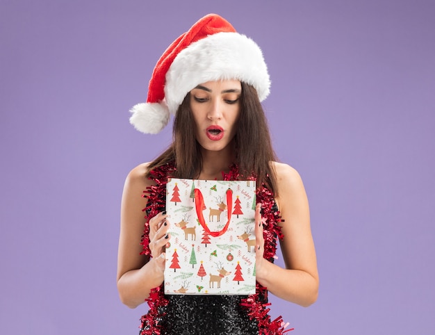 Hermosa joven sorprendida con sombrero de navidad con guirnalda en el cuello sosteniendo y mirando una bolsa de regalo aislada sobre fondo púrpura