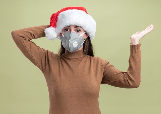 Hermosa joven sorprendida con sombrero de navidad con puntos de máscara médica con la mano al lado poniendo la mano en la cabeza aislada sobre fondo verde oliva