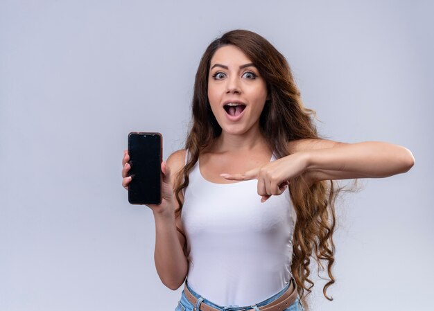 Hermosa joven sorprendida sosteniendo el teléfono móvil y apuntando a él en la pared naranja aislada con espacio de copia