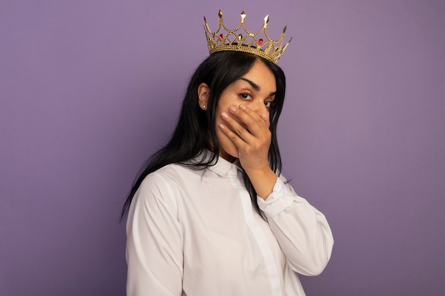 Hermosa joven sorprendida vestida con camiseta blanca y boca cubierta de corona con la mano aislada en púrpura