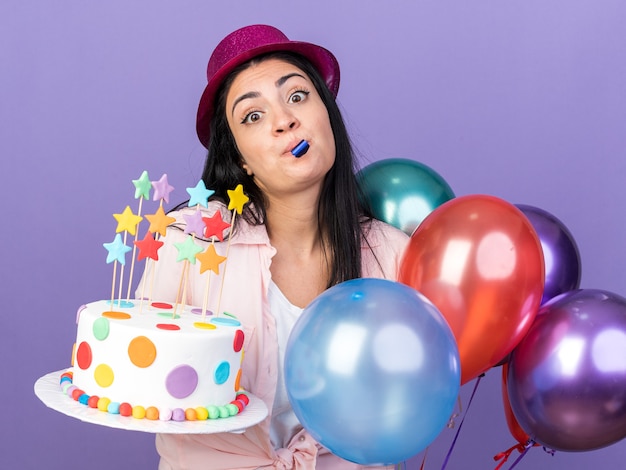 Hermosa joven sorprendida vistiendo gorro de fiesta de pie cerca de globos sosteniendo pastel soplando silbato de fiesta