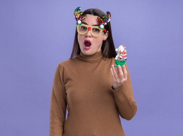 Hermosa joven sorprendida vistiendo un suéter marrón con gafas de Navidad sosteniendo un juguete aislado sobre fondo azul.