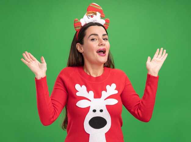 Hermosa joven sorprendida vistiendo un suéter de Navidad con aro de pelo de Navidad extendiendo las manos aisladas sobre fondo verde