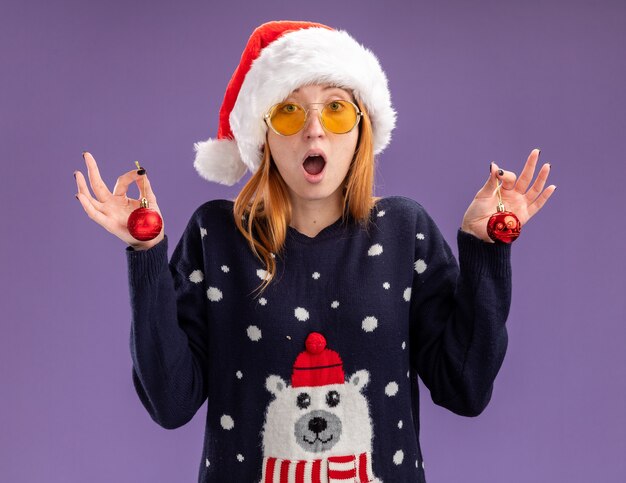 Hermosa joven sorprendida vistiendo un suéter de Navidad y un sombrero con gafas sosteniendo bolas de Navidad aisladas sobre fondo púrpura