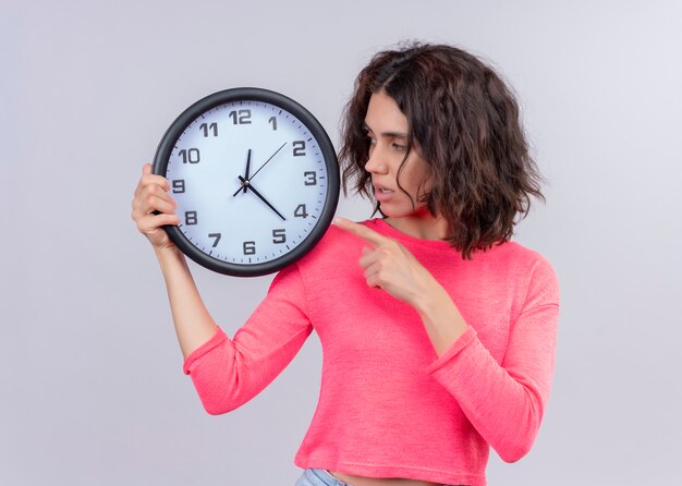 Hermosa joven sosteniendo el reloj y apuntando hacia él sobre fondo blanco aislado