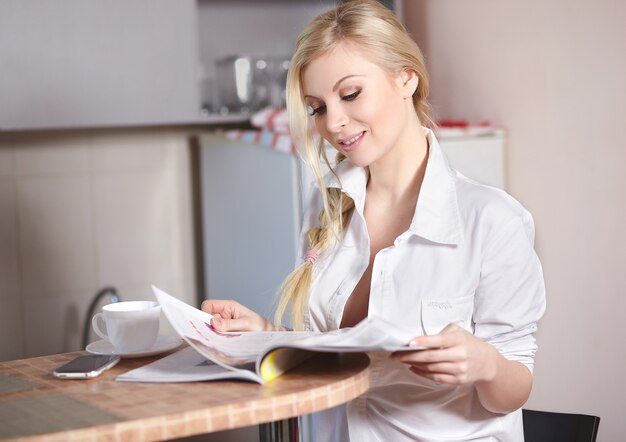 Hermosa joven sostiene sentado en la cocina y leer revista