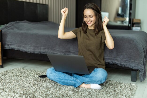 Hermosa joven tendida en el suelo usando una computadora portátil gritando orgullosa y celebrando la victoria y el éxito muy emocionada