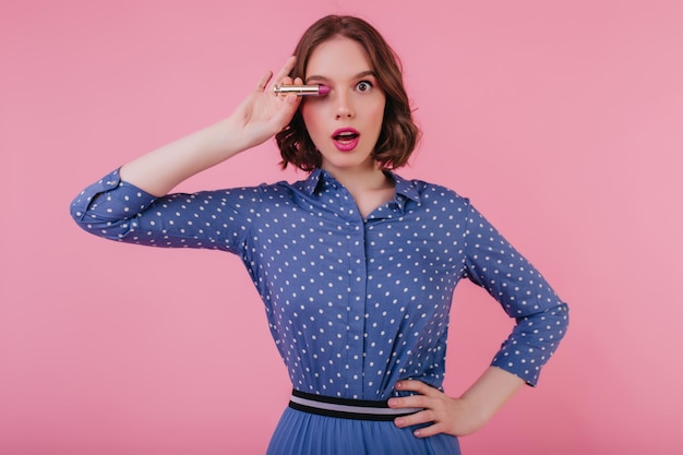 Foto gratuita hermosa joven en traje azul posando con expresión de sorpresa mientras se maquilla retrato de estudio de mujer de moda con cabello ondulado sosteniendo lápiz labial sobre fondo rosa