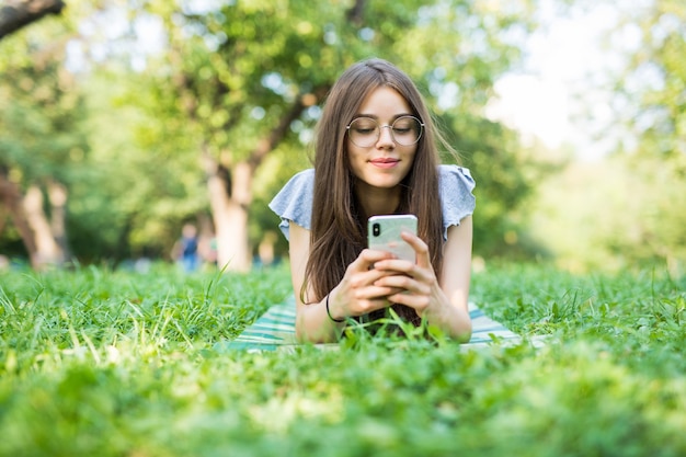 Hermosa joven tumbado en la hierba leyendo un mensaje en un teléfono celular en el parque