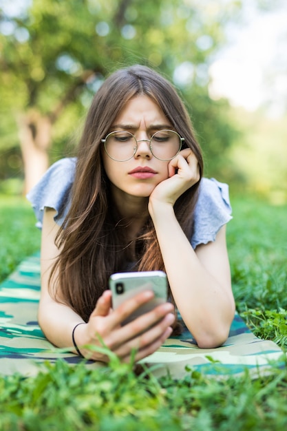 Hermosa joven tumbado en la hierba leyendo un mensaje en un teléfono celular en el parque