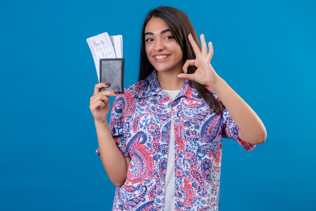 Foto gratuita hermosa joven turista sosteniendo pasaporte con boletos mirando a la cámara sonriendo alegremente haciendo bien firmar de pie sobre fondo azul aislado