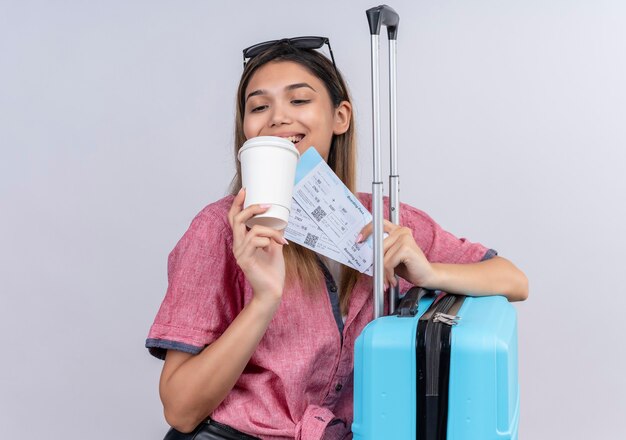 Una hermosa joven vestida con camisa roja y gafas de sol mirando una taza de café de plástico mientras sostiene boletos de avión y maleta azul sobre una pared blanca