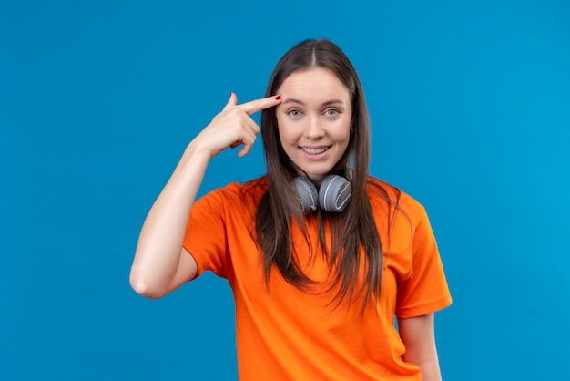 Hermosa joven vestida con camiseta naranja con auriculares apuntando templo recordándose a sí misma no olvidar lo importante sonriendo de pie sobre fondo azul aislado