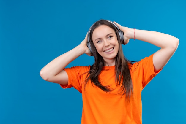 Hermosa joven vestida con camiseta naranja con auriculares disfrutando de su música favorita sonriendo feliz y positivo de pie sobre fondo azul aislado