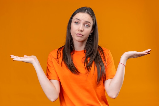 Hermosa joven vestida con camiseta naranja despistada y confundida encogiéndose de hombros extendiendo las manos sin respuesta de pie sobre fondo naranja aislado