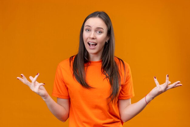 Hermosa joven vestida con camiseta naranja extendiendo los brazos mirando confundido como pregunta de pie sobre fondo naranja aislado