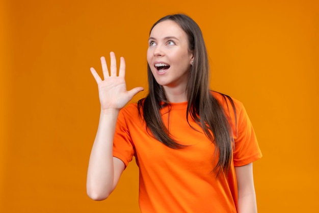 Hermosa joven vestida con camiseta naranja gritando o llamando a alguien con la mano cerca de la boca de pie sobre fondo naranja aislado