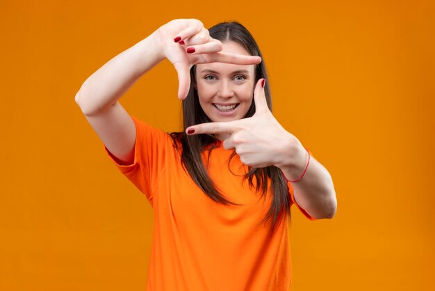 Hermosa joven vestida con camiseta naranja haciendo marco con las manos y los dedos sonriendo alegremente de pie sobre fondo naranja aislado
