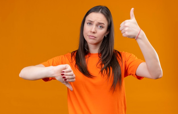 Foto gratuita hermosa joven vestida con camiseta naranja mirando a la cámara con expresión confusa mostrando los pulgares hacia arriba y hacia abajo de pie sobre fondo naranja aislado