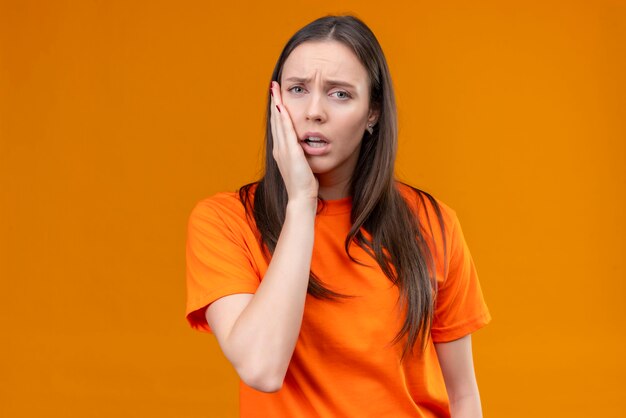 Hermosa joven vestida con camiseta naranja mirando mal tocando su mejilla sintiendo dolor de muelas de pie sobre fondo naranja aislado