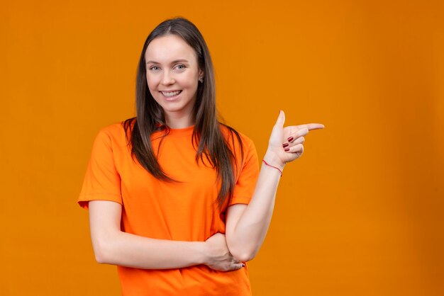 Hermosa joven vestida con camiseta naranja sonriendo positivo y feliz apuntando con el dedo hacia el lado de pie sobre fondo naranja aislado