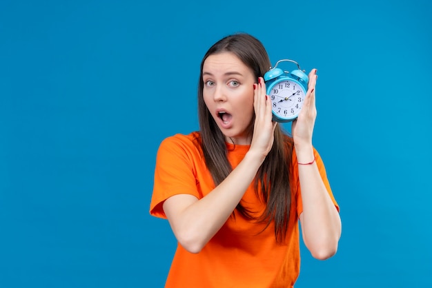 Hermosa joven vestida con camiseta naranja sosteniendo despertador mirando asombrado y sorprendido de pie sobre fondo azul aislado