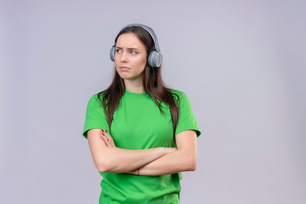 Hermosa joven vestida con camiseta verde con auriculares de pie con los brazos cruzados mirando a un lado disgustado sobre fondo blanco aislado