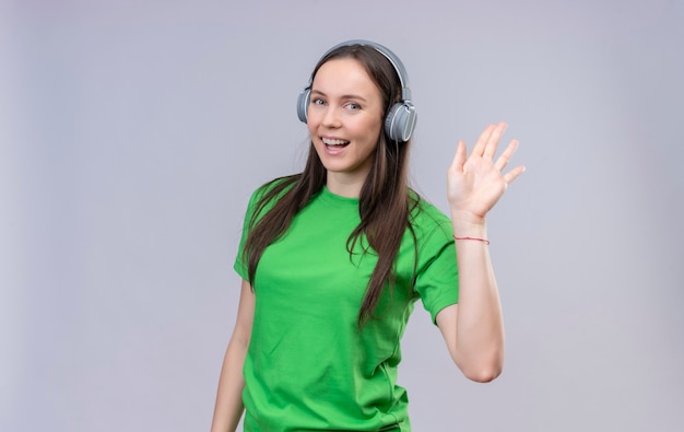 Hermosa joven vestida con camiseta verde con auriculares sonriendo alegremente saludando con la mano sobre fondo blanco aislado