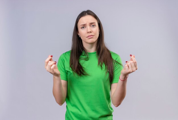 Hermosa joven vestida con camiseta verde mirando a la cámara disgustado frotándose los dedos haciendo gesto en efectivo pidiendo dinero de pie sobre fondo blanco aislado