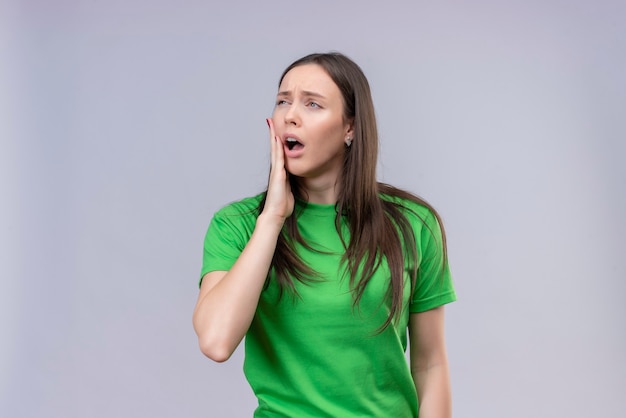 Hermosa joven vestida con camiseta verde mirando mal tocando su mejilla que sufre de dolor de muelas de pie sobre fondo blanco aislado