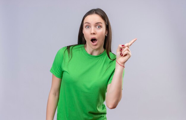 Hermosa joven vestida con camiseta verde mirando sorprendido y asombrado señalando con el dedo hacia el lado de pie sobre fondo blanco aislado