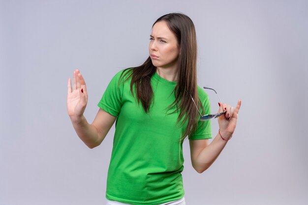 Hermosa joven vestida con camiseta verde de pie con los brazos levantados mirando confiado y satisfecho de sí mismo sobre fondo blanco aislado