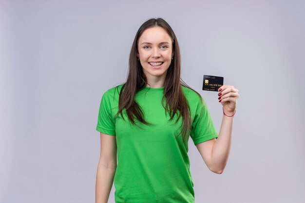 Hermosa joven vestida con camiseta verde positiva y feliz celebración de tarjeta de crédito sonriendo alegremente de pie sobre fondo blanco aislado