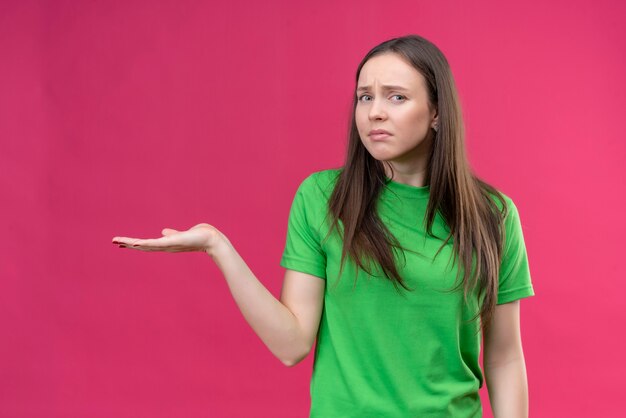 Hermosa joven vestida con camiseta verde que presenta espacio de copia con el brazo de su mano con cara infeliz expresión triste de pie sobre fondo rosa aislado