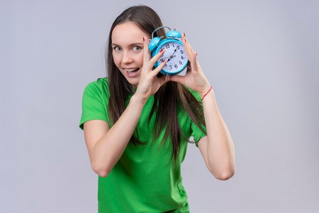 Hermosa joven vestida con camiseta verde con reloj despertador mirando a la cámara salió y feliz sonriendo de pie sobre fondo blanco aislado