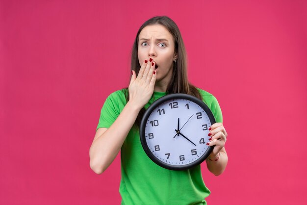 Hermosa joven vestida con camiseta verde con reloj mirando sorprendido y conmocionado cubriendo la boca con la mano sobre fondo rosa aislado