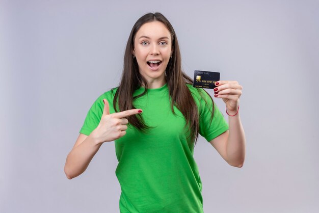 Hermosa joven vestida con camiseta verde salió y feliz celebración de tarjeta de crédito apuntando con el dedo a ella de pie sobre fondo blanco aislado