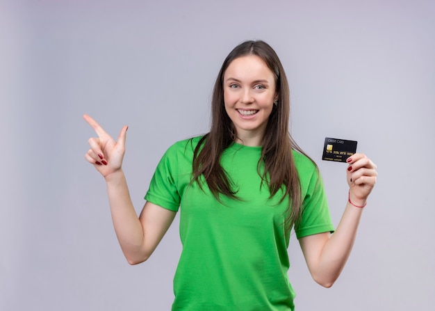 Foto gratuita hermosa joven vestida con camiseta verde salió y feliz celebración de tarjeta de crédito apuntando con el dedo hacia el lado de pie sobre fondo blanco aislado