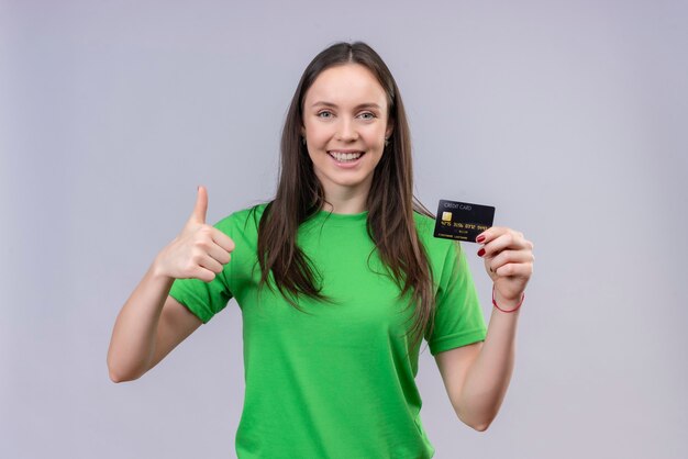 Hermosa joven vestida con camiseta verde salió y feliz celebración de tarjeta de crédito mostrando los pulgares para arriba sonriendo alegremente de pie sobre fondo blanco aislado