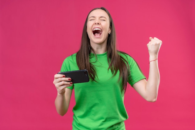 Hermosa joven vestida con camiseta verde con smartphone loco feliz levantando el puño cerrado sonriendo alegremente regocijándose de su éxito de pie sobre fondo rosa aislado