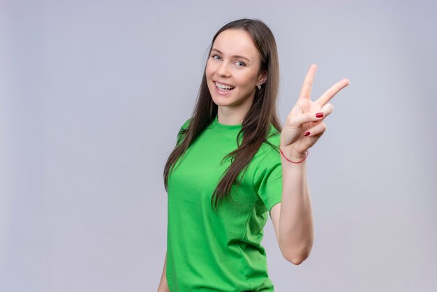 Hermosa joven vestida con camiseta verde sonriendo alegremente mostrando y apuntando hacia arriba con los dedos número dos o el signo de la victoria de pie sobre fondo blanco aislado