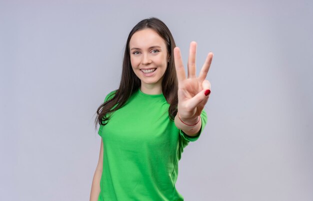 Hermosa joven vestida con camiseta verde sonriendo alegremente mostrando y apuntando hacia arriba con los dedos número tres de pie sobre fondo blanco aislado